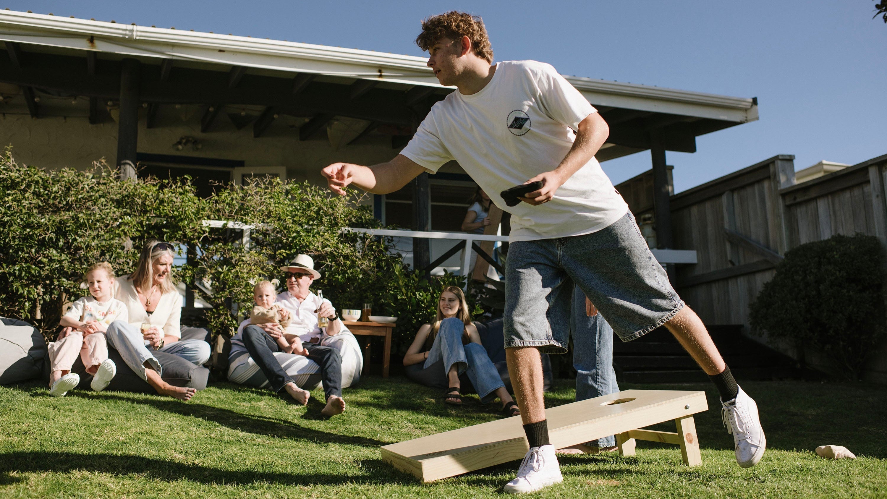 Guy throwing Cornhole Bag while friends watch in Backyard