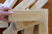Backyard Games Chats Tower wooden block with a humorous question printed on it, held over a pile of similar blocks.