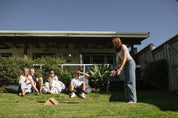 Group of people sitting on a grassy lawn in front of a house watching a girl play Backyard Games NZ Gutter Board. 